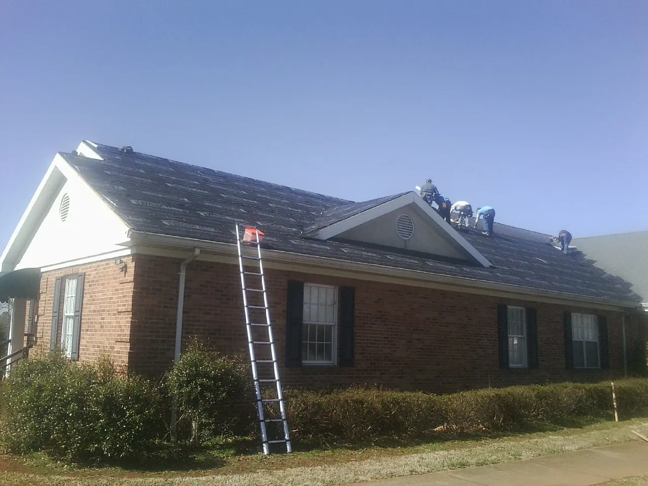 Crew tearing shingles off a brick ranch roof with a ladder set — Metal Roof Repair work by our crew in Argyle